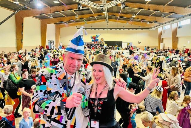 Moderatorin Jana am Mikro und DJ Merten schauen in die Kamera. Im Hintergrund machen kostümierte Kinder und Erwachsene eine Polonaise.
