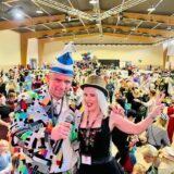 Moderatorin Jana am Mikro und DJ Merten schauen in die Kamera. Im Hintergrund machen kostümierte Kinder und Erwachsene eine Polonaise.