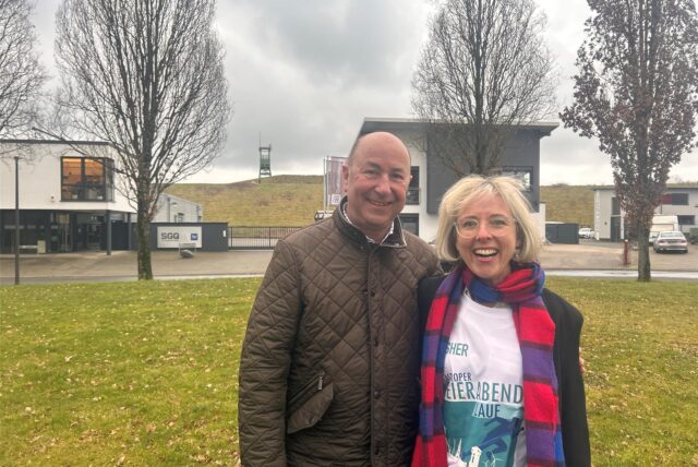 Wolfgang Heller und Anne Krüger stehen auf einer grünen Wiese, im Hintergrund ist die Zeche Erin.