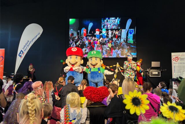 Die Comic-Helden Mario und Luigi von Paw Patrol stehen als Plüschfiguren auf der Bühne. Davor tanzen bunt kostümierte Kinder.
