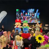 Die Comic-Helden Mario und Luigi von Paw Patrol stehen als Plüschfiguren auf der Bühne. Davor tanzen bunt kostümierte Kinder.