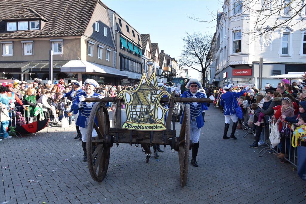 Beim Karnevalsumzug in Menden schieben Mitglieder der Mendener Karnevalsgesellschaft Kornblumenblau einen alten Wagen durch die Stadt. Links und rechts säumen hunderte Menschen die Straße Beim Karnevalsumzug in Menden schieben Mitglieder der Mendener Karnevalsgesellschaft Kornblumenblau einen alten Wagen durch die Stadt. Links und rechts säumen hunderte Menschen die Straße