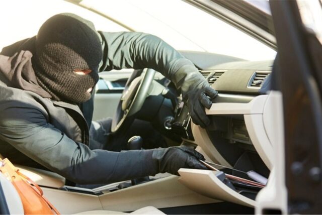 Symbolbild zeigt einen Einbrecher mit Skimaske, der das Handschuhfach eines Autos leerräumt.