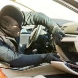 Symbolbild zeigt einen Einbrecher mit Skimaske, der das Handschuhfach eines Autos leerräumt.