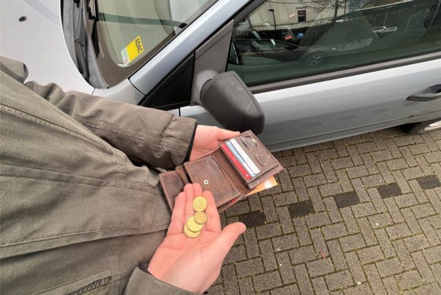 Ein Mann steht neben einem Auto, hält seine Geldbörse in der linken Hand und einige Münzen in der anderen. (Symbolbild)