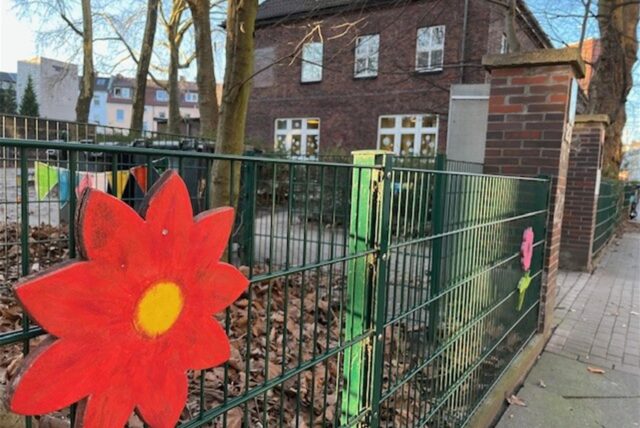 Am Zaun eines Kindergartengeländes hängt eine bunte Holzblume. Im Hintergrund steht ein Backsteingebäude.