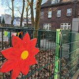 Am Zaun eines Kindergartengeländes hängt eine bunte Holzblume. Im Hintergrund steht ein Backsteingebäude.