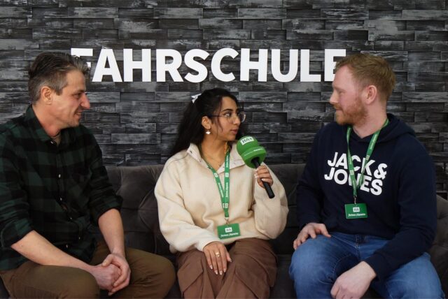 Fahrlehrer Sebahattin Altunsoy sowie Rosi Taslima Azam und Jonas Alder von der Hertener Allgemeinen sitzen bei einem Interview auf einem Sofa, im Hintergund ist „Fahrschule“ zu lesen.