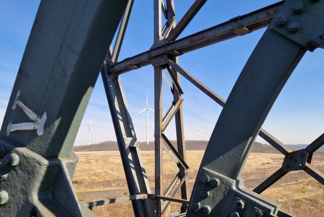 Ein Blick durch das Gerüst des Förderturms Auguste Victoria AV1 auf die Halde Brinkfortsheide in Marl. Zwischen den Stahlstreben sieht man die drei Windräder auf der Halde.