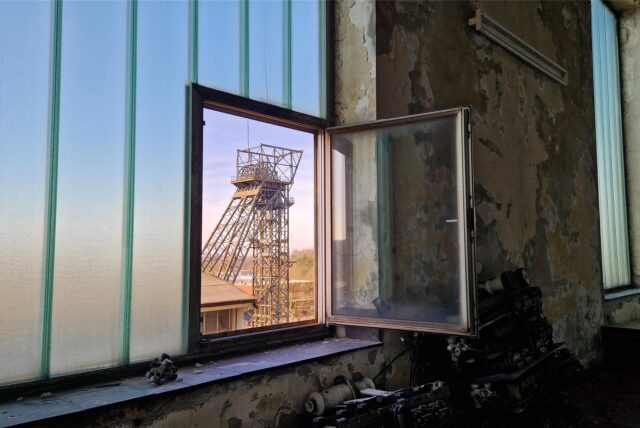 Das Bild zeigt ein Milchglasfenster aus schrägem Winkel. In diesem Milchglasfenster ist ein kleineres Fenster mit klarem Glas. Es steht offen. Der Blick durch das offene Fenster fällt auf den Förderturm Auguste Victoria 1 in Marl. Der Himmel ist Blau.