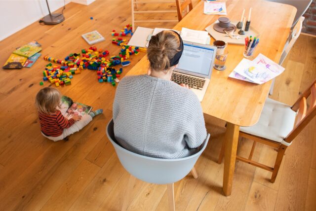 Mutter sitzt am Laptop und ein Kind spielt neben ihr auf dem Boden