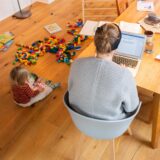 Mutter sitzt am Laptop und ein Kind spielt neben ihr auf dem Boden