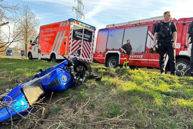 Einsatzfahrzeuge von Feuerwehr und Rettungsdienst stehen am Rand einer Straße, im Graben liegt ein blaues Motorrad. Zwei Polizisten stehen neben dem Graben.