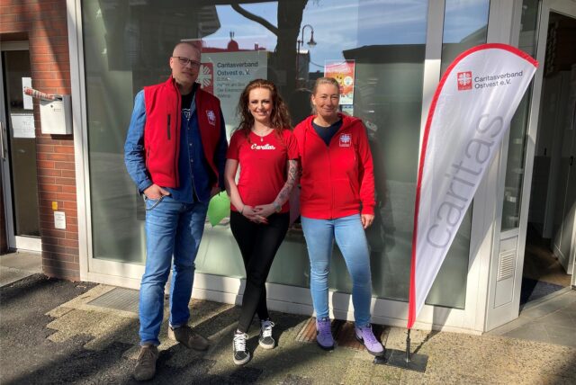 Eine Frau in roter Jacke steht neben einem Aufsteller mit der Aufschrift Caritas in der Fußgängerzone von Datteln.