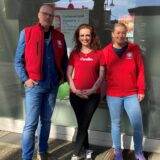 Eine Frau in roter Jacke steht neben einem Aufsteller mit der Aufschrift Caritas in der Fußgängerzone von Datteln.