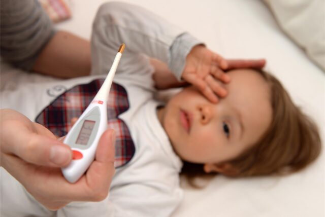 Ein Kind liegt im Bett und hält sich den Kopf und im Vordergrund hält eine Person ein Fieberthermometer.