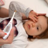 Ein Kind liegt im Bett und hält sich den Kopf und im Vordergrund hält eine Person ein Fieberthermometer.