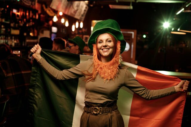 Eine Frau steht am St. Patrick’s Day in einer Bar und hält eine irische Flagge hoch.