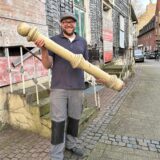 Ein Mann hält einen Pfeiler in der Hand. Er hat eine Kappe auf dem Kopf und steht vor einem alten Haus.