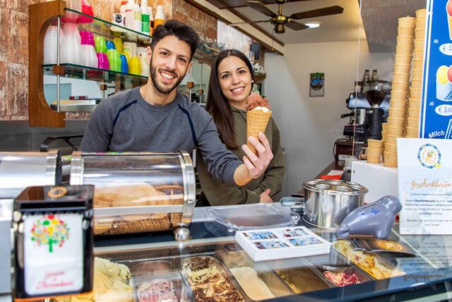 Miri Finocchiaro und Filippo Di Paola stehen im Eiscafé L’isola del Gelato in Herten-Westerholt und reichen ein Eis über den Verkaufstresen.