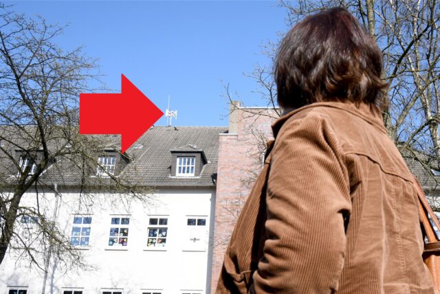 Eine Frau guckt auf eine Sirene auf einem Dach in Marl, die mit einem roten Pfeil gekennzeichnet ist.