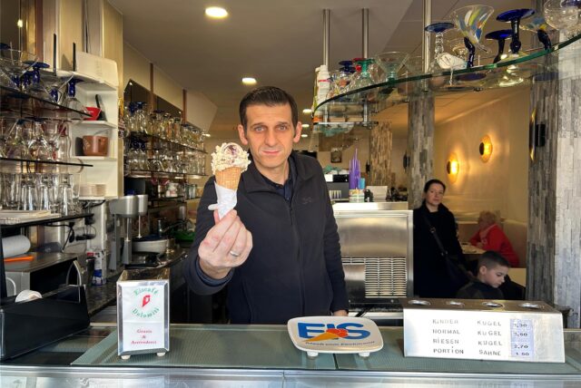 Ein Mann steht hinter einer Eistheke und hält eine Kugel Eis in einer Waffel in den Fingern.