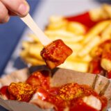 Eine Person piekst mit einer Holzgabel ein Stück Currywurst auf.