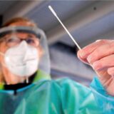 Eine Frau hat ein Abstrich-Stäbchen in der Hand, trägt einen grünen Kittel, Handschuhe, eine Maske und ein Visier.