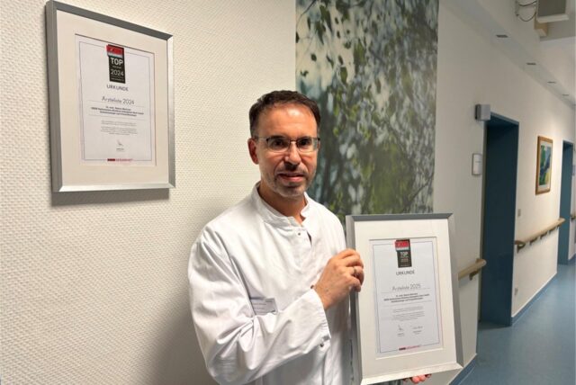 Dr. Ramon Martinez hat eine Urkunde in der Hand und steht vor einer weißen Wand