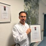 Dr. Ramon Martinez hat eine Urkunde in der Hand und steht vor einer weißen Wand