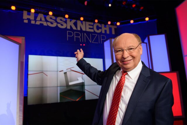 Hans-Joachim Heist alias Gernot Hassknecht steht in einem Studio und zeigt auf die Aufschrift „Hassknecht-Prinzip“.