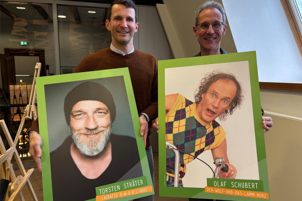 Marius Ebel und Lars Tottmann (r.) von der Arena Recklinghausen GmbH zeigen Fotos des diesjährigen Hurz-Preisträgers Olaf Schubert und seines Laudators Torsten Sträter.