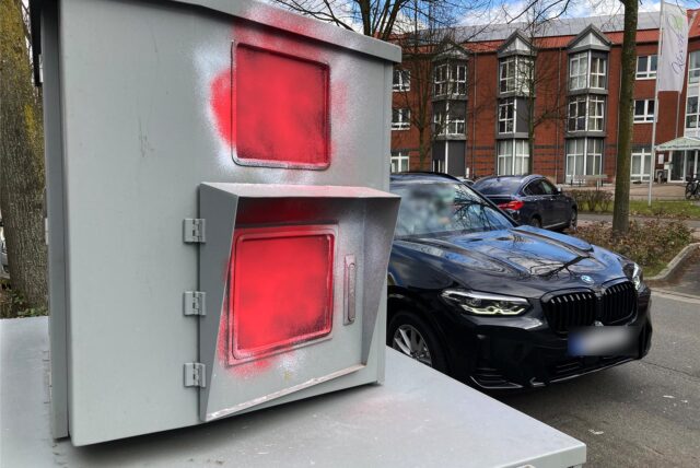 Unbekannte haben den Panzerblitzer an der Scherlebecker Straße in Herten mit roter Farbe beschmiert. Ein schwarzer BMW fährt in diesem Moment vorbei.