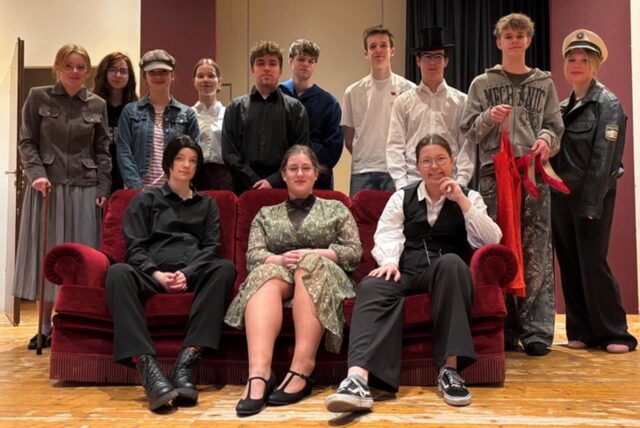 Eine Gruppe Schüler befindet sich auf einer Bühne, wovon drei Schüler auf einer roten Cordcouch sitzen und der Rest dahinter steht.