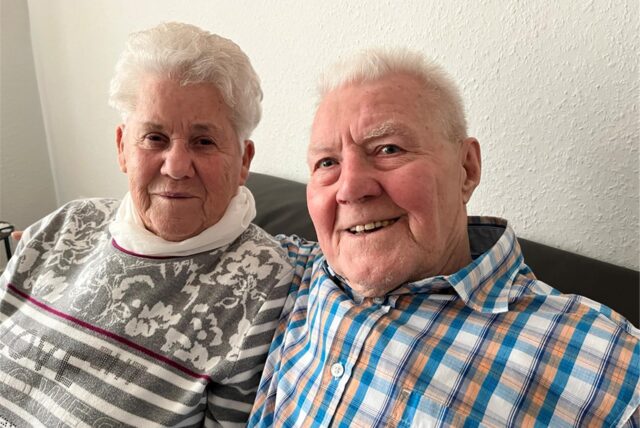 Renate und Josef Thesing sitzen auf der Couch und blicken in die Kamera.
