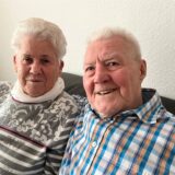 Renate und Josef Thesing sitzen auf der Couch und blicken in die Kamera.