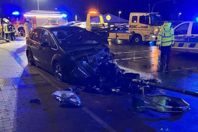 Ein Auto mit vollkommen zerstörter Front steht auf eine regennassen Fahrbahn, dahinter ein Abschleppfahrzeug und Blaulicht.