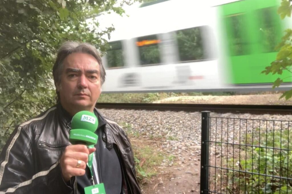 MZ-Redakteur Patrick Köllner mit einem Mikrofon, dahinter fährt ein Zug vorbei.