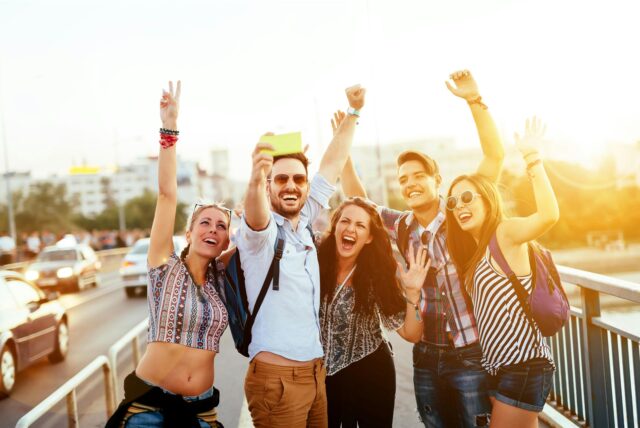 Fünf junge Erwachsene stehen auf einer Brücke und machen ein Selfie, während sie jubelnd in die Kamera lachen.