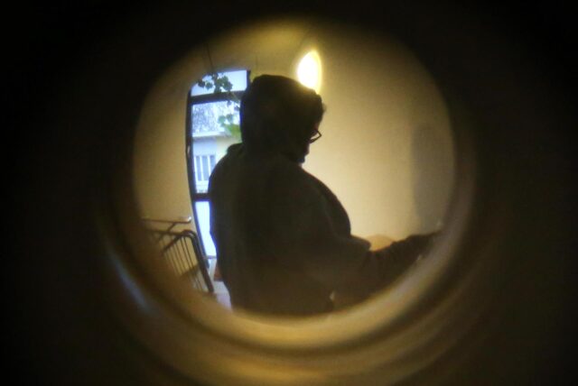 Das Symbolbild zeigt den Blick durch einen Türspion. Vor der Tür steht eine dunkel gekleidete Person, die nicht genauer zu erkennen ist.