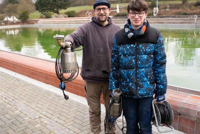 Schwimmmeister Kai Weinhardt und Azubi Thorben stehen vor dem mit grünem Wasser gefüllten 25-Meter-Becken im Copa Ca Backum.