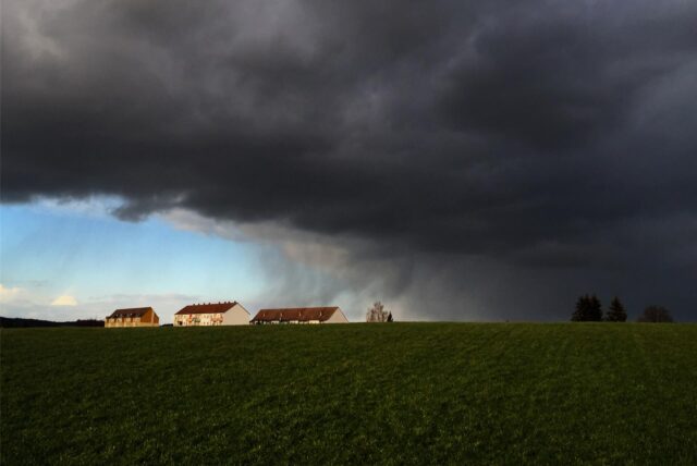 Dunkle Regenwolken ziehen über Häuser hinweg, während im Hintergrund blauer Himmel zu sehen ist.