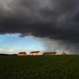 Dunkle Regenwolken ziehen über Häuser hinweg, während im Hintergrund blauer Himmel zu sehen ist.