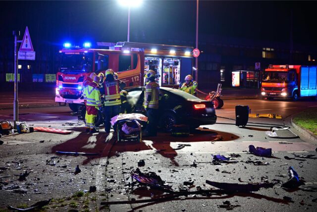 Feuerwehrleute an einem Fahrzeugwrack. Auf der Straße liegen Trümmerteile, im Hintergrund Feuerwehrfahrzeuge mit Blaulicht.