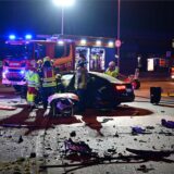 Feuerwehrleute an einem Fahrzeugwrack. Auf der Straße liegen Trümmerteile, im Hintergrund Feuerwehrfahrzeuge mit Blaulicht.