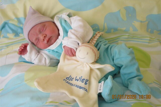 Svea liegt vor einem grün und blauen Hintergrund. Auf ihr liegt ein Kuscheltier auf welchem „Wiege, St. Rochus Hospital“ steht.