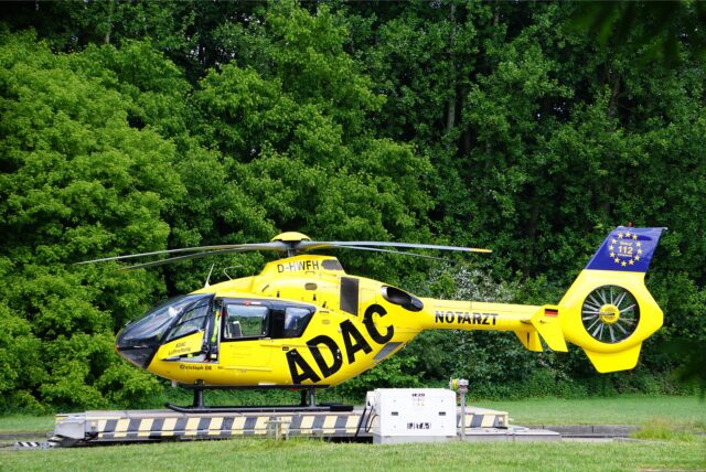 Ein gelber ADAC-Rettungshubschrauber mit der Aufschrift Notarzt steht auf einer Landeplattform auf einer Wiese. Im Hintergrund Bäume und dichtes Gesbüsch.
