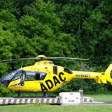 Ein gelber ADAC-Rettungshubschrauber mit der Aufschrift Notarzt steht auf einer Landeplattform auf einer Wiese. Im Hintergrund Bäume und dichtes Gesbüsch.