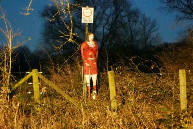 Eine Frauenfigur mit einem roten Kleid steht an einem Feldweg.