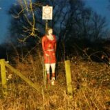 Eine Frauenfigur mit einem roten Kleid steht an einem Feldweg.